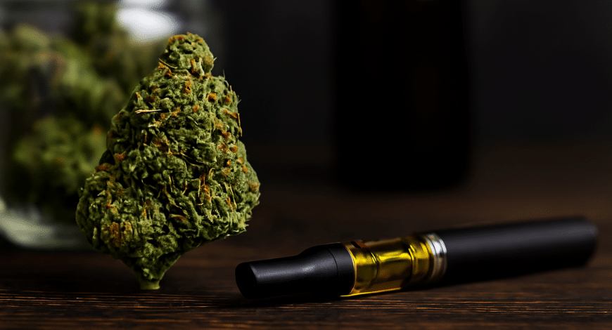 A dense indica cannabis bud next to a vape pen filled with oil, with a jar of buds blurred in the background on a wooden surface