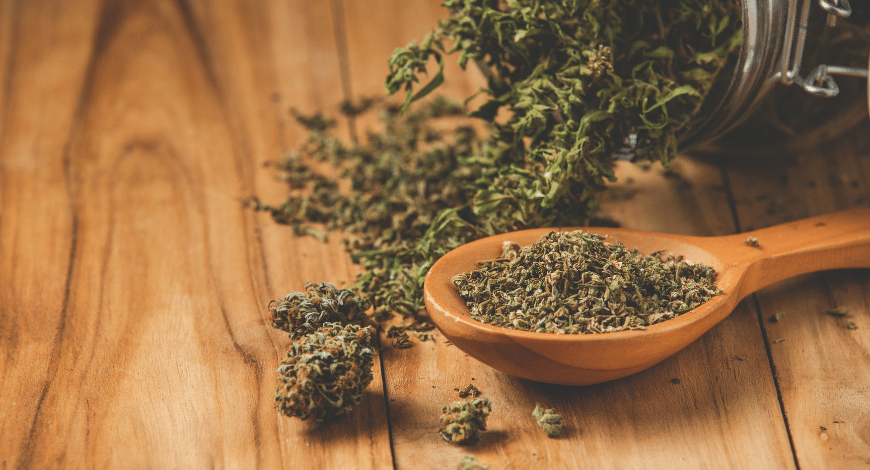 Loose and ground cannabis buds on a wooden surface with a jar and wooden spoon, representing a sativa strain